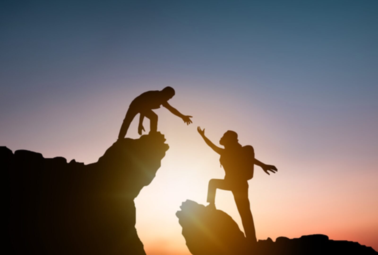 One person reaching down to help another up a rock face at sunset.